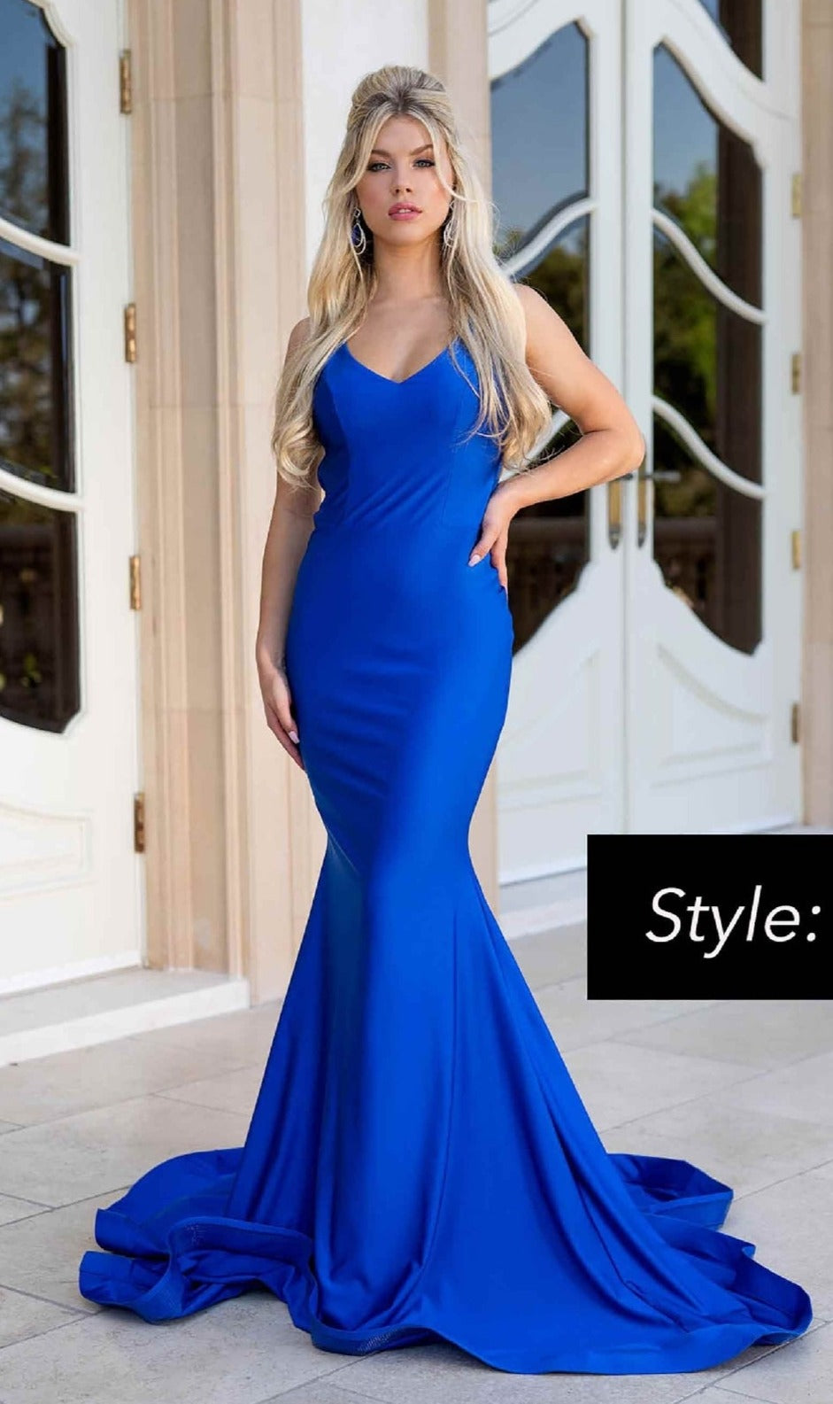 A woman stands outside in front of white double doors, wearing the Jessica Angel 635 Strappy-Back Long Prom Dress, a fitted royal blue floor-length gown with thin straps and a dramatic mermaid skirt.